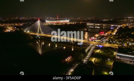 Luftansicht von Warschau bei Nacht, die eine beleuchtete Hängebrücke über einem dunklen Fluss, eine Uferpromenade, ein leuchtendes Stadion und eine belebte Ufergegend mit Reflexionen über dem modernen Stadtbild zeigt Stockfoto