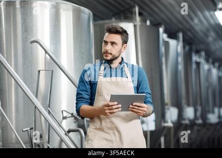 Junger kaukasischer Mann mit Schürze, der ein Tablet in einer Industrieküche hält, Geschäftskonzept Stockfoto