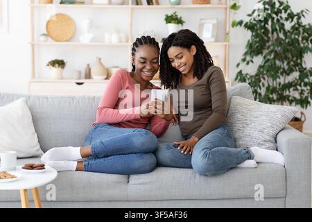 Fröhliche schwarze Freundin, die auf dem Sofa sitzt und das Smartphone benutzt Stockfoto