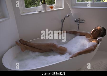 Mittelalte afroamerikanische Frau lehnt sich in der Badewanne zu Hause zurück mit Schaum, Handtuch-Kopfstütze, Pflanze Stockfoto