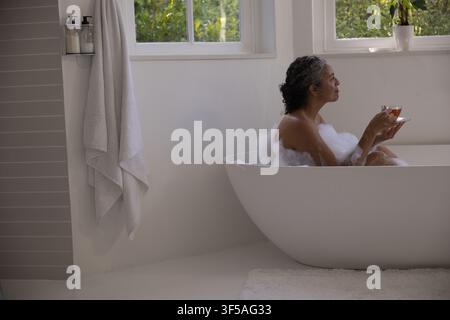 Ältere afrikanische Frau sitzt in einer schaumigen freistehenden Wanne im Badezimmer, hält einen Glasbecher, blickt Stockfoto