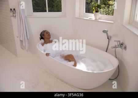 Afroamerikanische Seniorin liegt in einem schaumgefüllten ovalen Badewannenbecken im Badezimmer und ruht ihren Kopf auf einem Handtuch Stockfoto