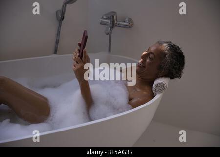 Senior Afroamerikanerin liegt in der Badewanne, hält das Telefon über den Schaum mit einem gerollten Handtuch Stockfoto