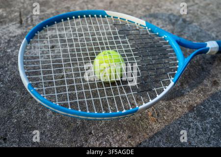 Nahaufnahme eines blauen Tennisschlägers, der auf rauem grauem Asphalt liegt, mit einem grünen Ball, der zentriert im Stringbett liegt, zeigt die Textur von Outdoor-Sport und Ausrüstungsdetails für Training, Übung, Wettkampf und Stockfoto