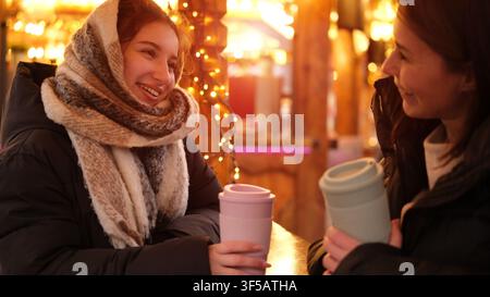 Junge Frauen genießen Gespräche und warme Getränke auf einem lebhaften Weihnachtsmarkt. Stockfoto
