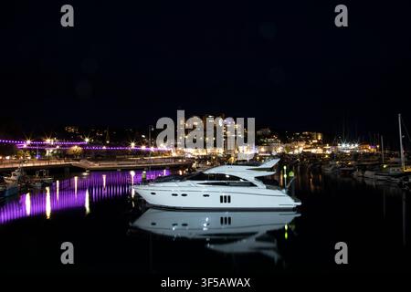 Ein luxuriöses Motorboot ruht auf ruhigem Wasser und wirft nachts ein scharfes Spiegelbild im Hafen von Torquay neben der beleuchteten Millennium Bridge. Stockfoto