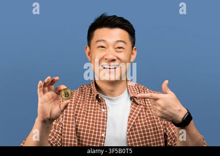 Lächelnder asiatischer Mann hält eine Bitcoin-Münze im Studio, Geschäftskonzept Stockfoto