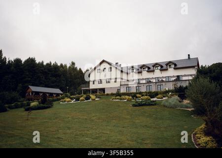 Rustikales Hotelgebäude mit Dachgauben mitten in einem üppigen grünen Rasen und Wald unter bewölktem Himmel. Gemütliche polnische Lodge, europäischer Landresort, Waldhütte-Urlaub, Eco-Reizziel Stockfoto