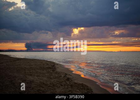 Dramatischer Sonnenuntergang über dem Ozean mit stürmischen Wolken und Rauch von einem fernen Feuer. Konzept von Klimawandel, Naturkatastrophe, dramatischem Küstenwetter, ökologischer Krise und intensiver Naturgewalt Stockfoto