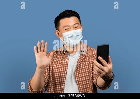 Zufriedener asiatischer Mann mit Gesichtsmaske bei einem Videoanruf, lächelnd vor blauem Hintergrund, Gesundheitskonzept Stockfoto
