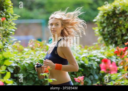 Eine blonde Frau in Sportkleidung läuft an blühenden Rosenbüschen vorbei mit einem Fitness-Drink in einem gestalteten Garten in Primrose Hill, London am 11. Juni 201 Stockfoto