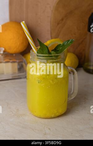 Eine vertikale Aufnahme eines erfrischenden gelben Zitrus-Orangen-Smoothies oder einer Limonade, serviert in einem gläsernen Mason-Jar-Becher mit einem Strohhalm, Minze und frischen Früchten. Stockfoto
