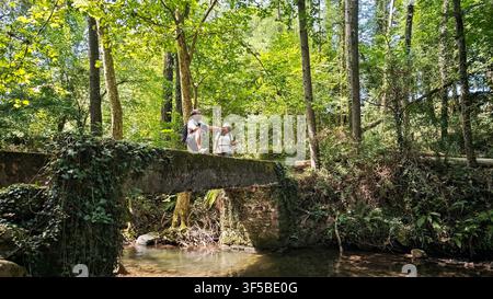 Zwei Reisende mit Rucksäcken stehen auf einer alten Betonfußgängerbrücke über einem Bach. Dichter grüner Wald in Erratzu, Baztan-Tal, Navarra, Spanien. Stockfoto