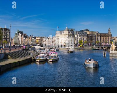 Amsterdam, Niederlande – 16. Apr 2026: Tourboote und ein kleines Vergnügungsschiff kreuzen den Damrak neben großen historischen Gebäuden im Zentrum von Amsterdam, Niederlande, unter einem strahlend blauen Himmel Stockfoto