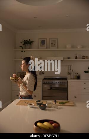 Afrikanische Frau, die eine Mahlzeit probiert, steht an der Kücheninsel, trägt ein gestreiftes Oberteil, hält eine Schüssel und eine Gabel Stockfoto