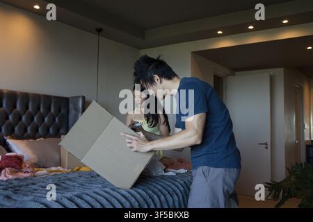 Asiatisches Paar packt auf dem Bett im Schlafzimmer aus, Mann in dunkelblauer Hemd handelt Kiste, Frau überprüft Smartphone Stockfoto