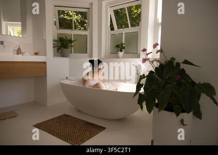 Afroamerikanische Frau badet in einer weißen Wanne im Badezimmer nahe der Fenster mit Schaum, hölzerner Waschtisch Stockfoto