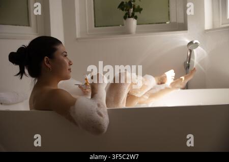 Afroamerikanische Frau lehnt sich in einer mit Seifenblasen gefüllten Badewanne zurück und hält ein Stielglas Stockfoto