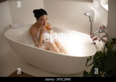 Afrikanische Frau sitzt in freistehender ovaler Badewanne im Badezimmer und nippt an einer klaren Teetasse aus Glas Stockfoto