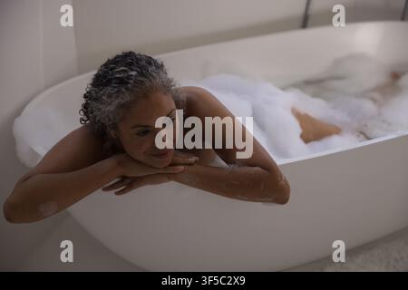 Afroamerikanische Seniorin lehnt sich in freistehender Badewanne zurück, das Kinn auf den Händen ruhend zwischen Blasen Stockfoto