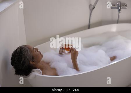 Senior afrikanisch-amerikanische Frau ruht in der Hausbadewanne mit Schaum, hält eine gläserne Teetasse und Untertasse Stockfoto