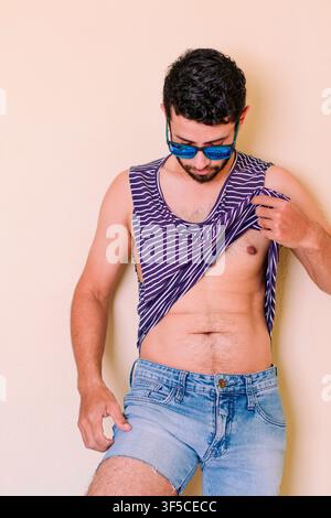 Junger Mann mit Sonnenbrille hebt sein Hemd und spürt die Hitze. Mann in Sommerkleidung, Fischer trägt einen Strohhut. Junger Mann in einem blauen Stockfoto