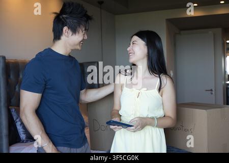 Asiatisches Paar packt Spendenkiste im Schlafzimmer, lächelnd, überprüft Telefon in Marineblauem T-Shirt, gelbem Kleid Stockfoto