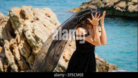 Junge Frau in schwarzem Kleid mit schwarzem Schal, die auf den Felsen steht Stockfoto