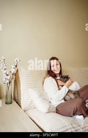 Glückliche Frau in einem weißen Strickpullover sitzt auf einem weichen beigen Bett und umarmt sanft ihre flauschige graue Katze mit einer Vase mit Baumwollblumen in der Nähe in einem warmen hellen Raum Stockfoto