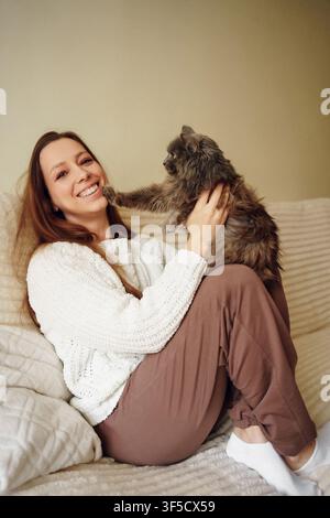 Ein warmes und intimes Porträt einer fröhlichen jungen Frau mit langen Haaren, die bequem auf einer weichen Couch zu Hause sitzt. Sie trägt einen strukturierten cremeweißen Pullover und braune Lounge-Hosen und ist glücklich Stockfoto