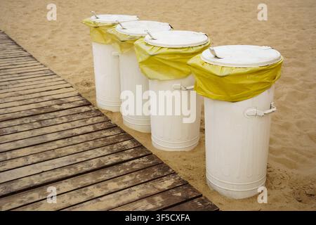 Vier weiße Mülltonnen mit gelben Müllsäcken, die auf einem sandigen Strand platziert sind, Strandmüllmanagement, Küstensanitationseinrichtungen, Sauberkeit öffentlicher Parks, umweltfreundliche Meeresküstenwartung Stockfoto