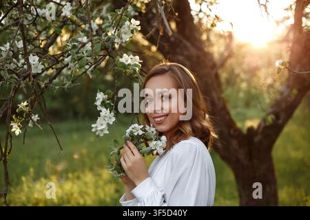 Junge Frau lächelt in einem blühenden Apfelgarten während der goldenen Stunde bei Sonnenuntergang, umgeben von weißen Blüten, Blumenlandschaft, sanftem Lebensstil, natürlichem Licht, ruhiger Gartenatmosphäre Stockfoto