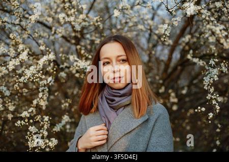 Junge Frau mit roten Haaren in einem grauen Wollmantel vor einem blühenden Kirschbaum stehend, ruhiges Frühlings-Lifestyle-Porträt, Outdoor-Mode, saisonale Natur-Schönheit und frische Blüte Stockfoto