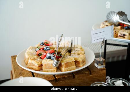 Scheiben Apfelkuchen mit Erdbeeren und Blaubeeren auf einem weißen Teller mit Servierzange, präsentiert für eine Catering-Veranstaltung, Dessertauswahl, Pastry-Catering, kulinarische Präsentation Stockfoto
