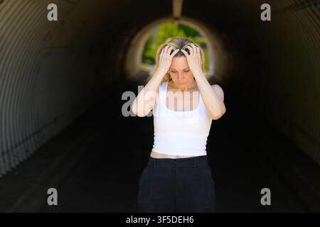 Emotionale Frau, die ihren Kopf in einem dunklen städtischen Tunnel hält, mit dramatischem natürlichem Gegenlicht und nachdenklichem Blick nach unten. Ausdrucksstarkes Lifestyle-Konzept Stockfoto
