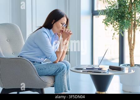 Nachdenkliche reife Geschäftsfrau sitzt in einem Sessel und starrt auf den Laptop-Bildschirm Stockfoto