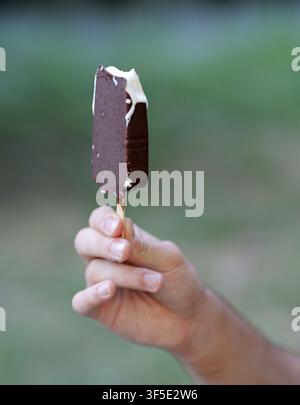 Hand, die eine köstliche, teilweise gegessene Schokoladen-Vanille-Eisriegel an einem Stock hält Stockfoto