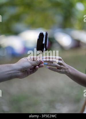 Zwei Hände, eine männliche und eine weibliche mit einem silbernen Ring und blau manikürten Nägeln, teilen einen schmelzenden schokoladenüberzogenen Vanilleeisstab auf einem Stiel du Stockfoto
