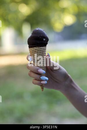 Person, die ein erfrischendes schokoladenüberzogenes Eis mit bunten Maniküren genießt, symbolisiert Sommer, Dessert und süße Vergnügungen im Freien Stockfoto