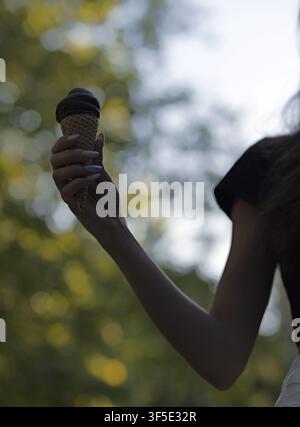 Eine Hand mit lackierten Nägeln hält eine Schokoladeneiswaffel vor einem verschwommenen Outdoor-Hintergrund und genießt eine süße Leckerei in der Natur bei warmem Wetter Stockfoto