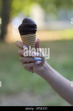 Frauenhänd mit Farbverlauf-Maniküre hält eine köstliche Schokoladeneiswaffel und genießt einen erfrischenden Snack bei warmem Sommerwetter im Freien Stockfoto