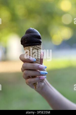 Frauenhand mit French Maniküre und blauen Nägeln, die ein köstliches schokoladenüberzogenes Eis hält und einen erfrischenden Snack an einem warmen, sonnigen Tag genießt Stockfoto