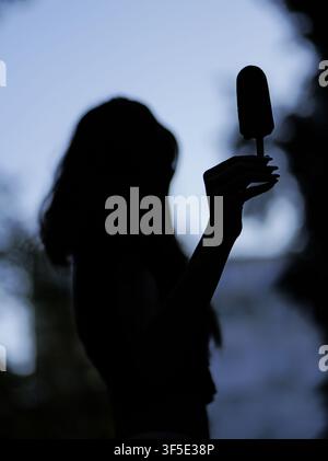 Frauensilhouette hält eine Eisbar gegen einen hellblauen Himmel, entspannt und genießt eine süße Leckerei im Freien während des Sommerabends Stockfoto