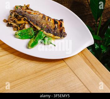 Gegrilltes Wolfsbarschfilet serviert mit sautierten Pilzen und Shishito-Pfeffern auf einem modernen weißen Teller Stockfoto