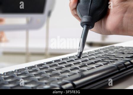 Eine Person verwendet eine manuelle schwarze Gummi-Luftblaspumpe, um Staub und Schmutz zwischen den Tasten einer Computertastatur in einer Büroumgebung zu entfernen Stockfoto