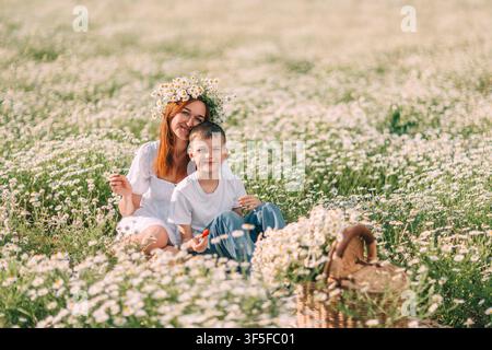 Eine Frau mit einem Blumenkranz sitzt mit ihrem Sohn in einem Feld weißer Gänseblümchen neben einem Weidenkorb. Stockfoto