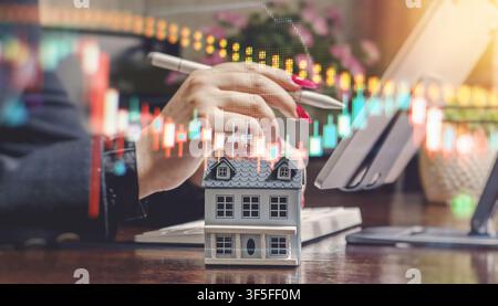 Konzept für Immobilienhandel und Finanzanalyse. Modellhaus mit Börendiagrammen und Person, die ein Tablet verwendet. Stockfoto