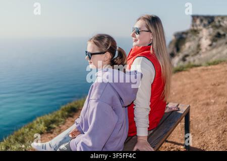 Mutter Tochter Klippenufer sitzend auf einer Holzbank mit Blick auf einen weiten blauen Ozean während eines malerischen Sommerurlaubs Stockfoto