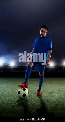 Ein junger indonesischer südostasiatischer Fußballspieler in einem blauen Trikot steht mit einem Fuß auf einem Fußball auf einem grünen Rasen in einem hell erleuchteten Sta Stockfoto
