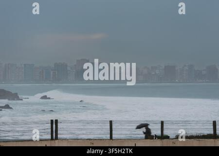 Mann mit Regenschirm geht während starkem Regen und Wind entlang eines Zauns. Skyline von Gijon im Hintergrund sichtbar durch den Nebel. Asturien, Spanien. Stockfoto